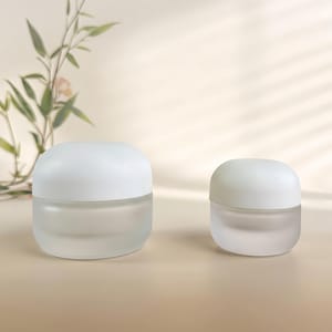 May include: Two round cosmetic jars with frosted glass bases and smooth white lids. The larger jar is positioned on the left, and the smaller jar is on the right. Both containers feature a simple, rounded silhouette with a clean, minimalist aesthetic.