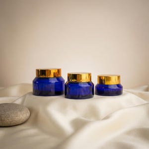 以下が含まれることがあります： 金色の蓋が付いたコバルトブルーのガラス瓶が3つ、白いサテンの表面に配置されています。滑らかな灰色の石がサテンの上に置かれています。瓶はサイズが異なり、化粧品や保管に適しています。