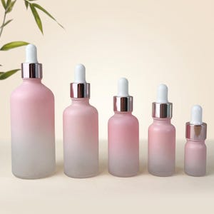 May include: Five glass dropper bottles arranged in a row from largest to smallest. Each bottle features a frosted finish with a pink to white gradient, a silver metallic collar, and a white rubber dropper top for dispensing liquids.