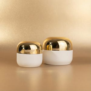 Pode incluir: Dois potes cosméticos brancos com tampas douradas arredondadas. Os potes estão sobre um fundo dourado. Os potes são de tamanhos diferentes, o maior à direita.