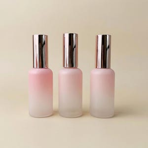 May include: Three glass spray bottles with a pink and white gradient. The bottles have silver-colored caps. The bottles are arranged in a row against a neutral background. The bottles are empty.