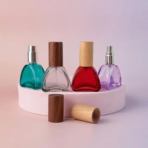 Puede incluir: Cuatro frascos de perfume de vidrio de colores con tapas de madera. Los frascos son triangulares y vienen en verde azulado, transparente, rojo y morado. Dos tapas de madera están separadas de los frascos. Los frascos se exhiben en una plataforma rosa redondeada.