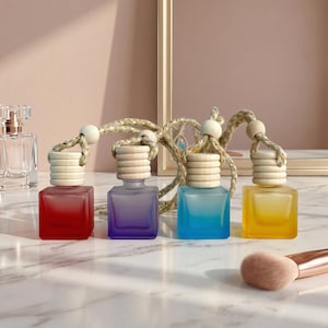 May include: Four colourful car air fresheners in red, purple, blue, and yellow. Each square glass bottle has a wooden cap and a rope for hanging. A makeup brush is in the foreground. The background includes a perfume bottle and a decorative frame.