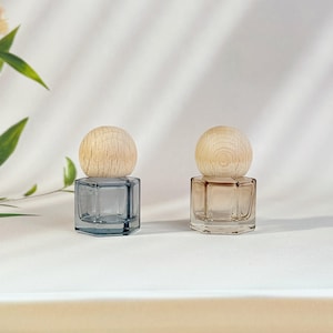May include: Two small, hexagonal glass bottles with wooden spherical stoppers. One bottle is a light blue-grey, the other is a light amber color. The bottles are displayed on a white surface with soft shadows.