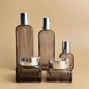Pode incluir: Uma coleção de frascos e potes de vidro castanho com tampas prateadas. O conjunto inclui dois frascos de spray, dois frascos quadrados e um frasco pequeno e arredondado. Os recipientes estão dispostos sobre um fundo neutro.