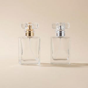 May include: Two clear glass perfume bottles with rectangular bodies and faceted, crystal-like stoppers. One has a gold-colored atomizer, and the other has a silver-colored atomizer. The bottles are displayed on a neutral background.