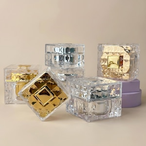 Pode incluir: Uma coleção de potes cosméticos quadrados de acrílico transparente com texturas facetadas semelhantes a diamantes. Os potes possuem tampas reflexivas em acabamentos dourado e prateado, alguns posicionados na vertical e um inclinado.