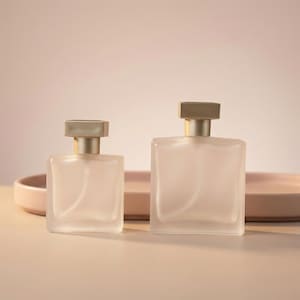 May include: Two frosted glass perfume bottles with gold-coloured caps. The bottles are rectangular and sit on a light pink tray. The background is a soft, neutral colour.