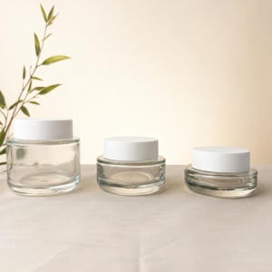 Puede incluir: Tres frascos de vidrio transparente de diferentes alturas, cada uno con una tapa circular blanca mate y lisa. Los frascos están dispuestos en fila, mostrando un diseño cilíndrico simple con bases de vidrio grueso para contener cremas.