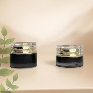 May include: Two cylindrical cosmetic jars with black glass bases and clear, faceted lids featuring gold-toned metallic accents. The containers are positioned side-by-side, showcasing a sleek, reflective design with a clear separation between the base and the lid.