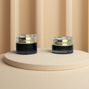 Op de afbeelding: Twee kleine, ronde, zwarte potjes met doorzichtige, facetgeslepen deksels en goudkleurige banden. De potjes worden getoond op een beige, cirkelvormig platform tegen een achtergrond van verticale, beige panelen. De potjes lijken voor cosmetica of huidverzorgingsproducten te zijn.