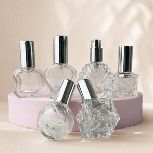 Könnte beinhalten: Eine Auswahl an transparenten Parfümflaschen aus Glas mit silberfarbenen Verschlüssen. Die Flaschen haben verschiedene Formen, darunter ein Herz und ein Apfel. Sie sind auf einer rosa Plattform vor neutralem Hintergrund angeordnet.