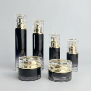 May include: An assortment of luxury cosmetic containers. The set includes several black and clear glass bottles and jars with gold-colored accents. The bottles have clear, textured tops. The jars have gold-colored lids.