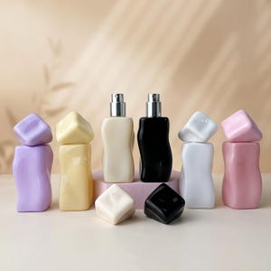 May include: A collection of six small, wavy-shaped perfume bottles with silver spray nozzles. The bottles come in lavender, yellow, cream, black, white, and pink, each featuring a matching removable square cap resting beside or atop the bottle.