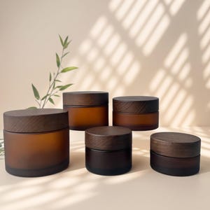 May include: A collection of five cylindrical jars with dark brown wooden lids and frosted amber glass bodies. The jars vary in size, arranged in a group with the largest on the left and smaller ones toward the right, featuring a smooth finish.
