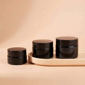 Puede incluir: Tres frascos cosméticos negros con tapas brillantes están colocados en una bandeja beige. Los frascos varían en tamaño, el más pequeño a la izquierda y el más grande a la derecha. El fondo es de un color suave y neutro.