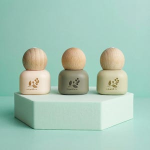 Może przedstawiać: Trzy małe, dekoracyjne butelki z drewnianymi zakrętkami. Butelki są w pastelowych kolorach: różowym, zielonym i żółtym. Każda butelka ma kwiatowy wzór i napis "-Luck-" z przodu. Butelki są wystawione na białym, geometrycznym stojaku.