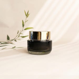 May include: A small, round, dark gray glass cosmetic jar with a reflective gold-toned metallic lid. The container has a smooth, cylindrical body and a flat base, designed for holding creams or other skincare products in a compact, elegant form.