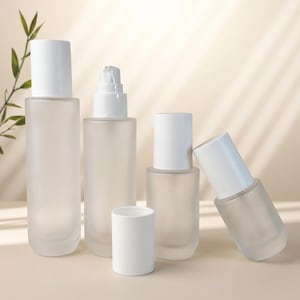 May include: A collection of four frosted glass cosmetic bottles with white caps and pumps. Three bottles are standing upright in varying heights, while one smaller bottle is tilted at an angle. A separate white cylindrical cap sits in the front.