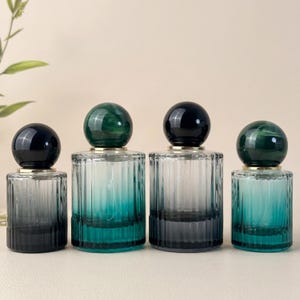 May include: Four glass perfume bottles with ribbed sides and spherical caps. The bottles have a gradient color scheme, transitioning from dark to teal. The caps are either dark green or black, with a gold-colored band at the base of the cap.