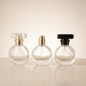 Op de afbeelding: Drie heldere glazen parfumflesjes met verschillende dopontwerpen. De ronde flesjes bevatten een heldere vloeistof en verstuivers. De doppen zijn goudkleurig, zwart en helder, met een facetontwerp op één. De flesjes staan op een lichtgekleurd oppervlak.