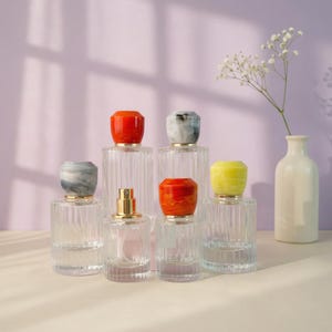 Puede incluir: Botellas de perfume de vidrio transparente con tapones coloridos. Los tapones son de color rojo, amarillo y gris. Las botellas están dispuestas sobre una superficie clara contra un fondo púrpura.