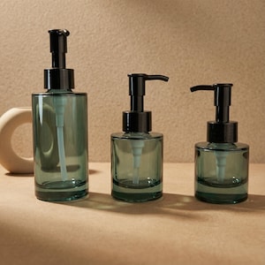 May include: Three glass soap or lotion dispensers in varying sizes. The dispensers are a muted teal color with black pump tops. The bottles are cylindrical and have a minimalist design, suitable for bathroom or kitchen use.