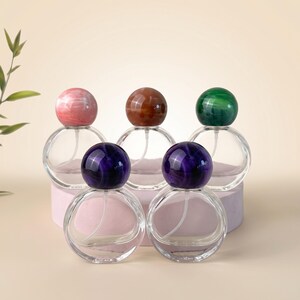 May include: Five clear glass perfume bottles with spherical bodies and round, marbled caps in pink, brown, green, and two shades of purple. Each bottle features a white internal spray tube and a smooth, transparent glass construction for liquid storage.