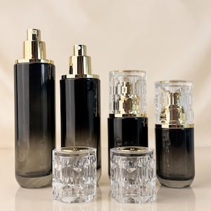 May include: An assortment of black and gold cosmetic bottles and jars. The bottles have a gradient effect, transitioning from black to clear. The jars are clear with a geometric design. The tops of the bottles and jars are gold.