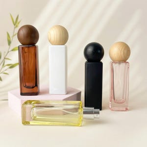 May include: An assortment of rectangular perfume bottles in various colors, including amber, white, black, and pink, each topped with a wooden sphere. One bottle lies horizontally, showcasing a yellow liquid and a silver atomizer. The bottles are arranged on a light-colored surface.