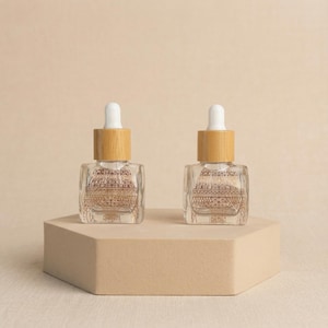 May include: Two clear glass dropper bottles with wooden caps and white droppers. The bottles have a gold Eiffel Tower design. They are displayed on a beige hexagonal platform against a neutral background.