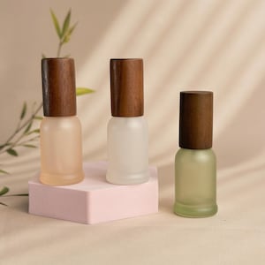 Könnte beinhalten: Drei Flaschen aus mattiertem Glas mit Holzverschlüssen. Die Flaschen sind in den Farbtönen Pfirsich, Weiß und Grün gehalten. Die Flaschen werden auf einem rosa geometrischen Block mit neutralem Hintergrund und einem Zweig Grünzeug präsentiert.
