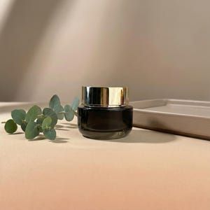 Op de afbeelding: Een kleine, donkere glazen pot met een goudkleurige deksel. De pot is rond en staat op een lichtgekleurd oppervlak. Een takje groene bladeren staat naast de pot. De achtergrond is een neutrale beige kleur.