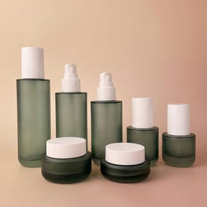 Peut inclure: Ensemble de contenants cosmétiques en verre dépoli avec couvercles blancs et distributeurs à pompe. L'ensemble comprend des flacons et des pots de différentes tailles, disposés sur un fond neutre. Les contenants sont de couleur vert sourd.