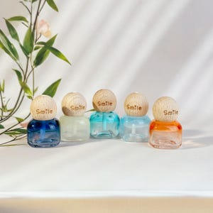 May include: Five small, colorful glass bottles with wooden caps. Each cap is engraved with the word "Smile." The bottles are filled with different colored liquids: blue, clear, turquoise, light blue, and orange. The bottles are arranged in a row.