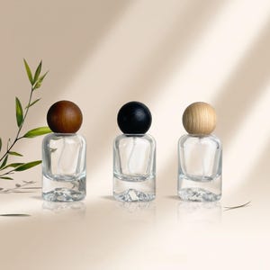 Puede incluir: Tres frascos de perfume de vidrio transparente con bases redondeadas y tapas esféricas de madera. Las tapas tienen acabados en tonos marrón oscuro, negro y madera natural clara. Cada frasco cuenta con un mecanismo de pulverización interno.