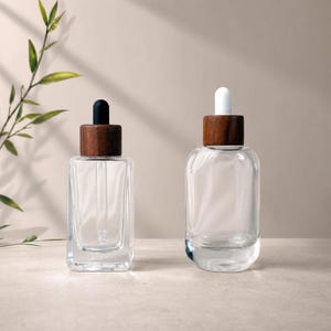 Könnte beinhalten: Zwei transparente Glas-Tropfflaschen mit Holzverschlüssen. Eine Flasche ist rechteckig, die andere abgerundet. Die Tropfer sind schwarz und weiß. Die Flaschen sind leer und stehen auf einer neutralen Oberfläche.