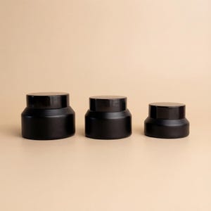 Pode incluir: Três potes cosméticos pretos com tampas pretas combinando estão alinhados. Os potes são de tamanhos diferentes, com um acabamento fosco. O fundo é de uma cor neutra.