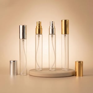 Puede incluir: Atomizadores de perfume de vidrio transparente con tapas y pulverizadores plateados y dorados. Los atomizadores se exhiben en una plataforma circular beige. Una tapa plateada está separada.