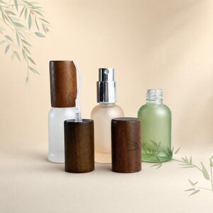 May include: Three small glass bottles with cylindrical wooden caps. One bottle is frosted clear, one is frosted light green, and one is clear with a silver spray pump mechanism. The wooden caps are dark brown with a smooth, cylindrical finish.