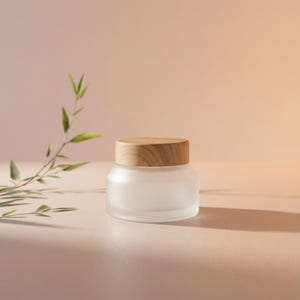 Op de afbeelding: Een matglazen pot met een licht houten deksel. De pot is rond en staat op een lichtroze oppervlak. Een takje met groene bladeren staat op de achtergrond. De pot is waarschijnlijk voor huidverzorgingsproducten.