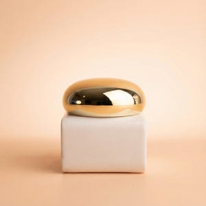 Pode incluir: Um objeto dourado em forma oval com uma superfície refletora está em cima de um bloco retangular branco. O objeto tem uma aparência lisa e polida, e o fundo é de cor pêssego suave.