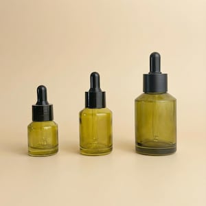 May include: Three olive-green glass dropper bottles with black tops. The bottles are different sizes, arranged from smallest to largest. The background is a solid, light beige color.