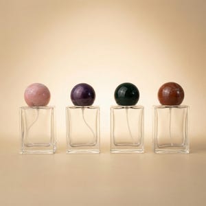 Puede incluir: Cuatro frascos de perfume de vidrio transparente con tapones esféricos de piedras preciosas en rosa, morado, verde y marrón. Los frascos son rectangulares con un pulverizador y un tubo curvo. El fondo es de color beige claro.