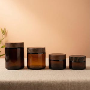 Puede incluir: Cuatro tarros de vidrio ámbar con tapas de madera de diferentes tamaños están dispuestos sobre una superficie neutra. Los tarros tienen forma cilíndrica y una estética cálida. Las tapas de madera contrastan con el vidrio.