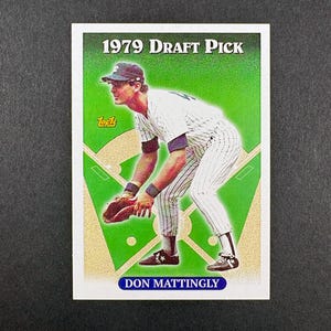 May include: A baseball card featuring Don Mattingly in a New York Yankees uniform. The card has a green background with the text "1979 Draft Pick" at the top and "Don Mattingly" at the bottom. The Topps logo is visible.