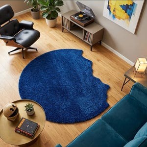May include: A circular, deep blue shag rug with a distinct bite-shaped cutout along one edge, featuring a thick, textured pile. The rug is positioned on a light-colored wooden floor in a room containing a black lounge chair, a record player console, and a gold-toned coffee table.