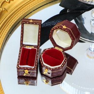 May include: Two open, burgundy leather jewelry boxes with gold accents. One is rectangular, the other is octagonal. Both have red velvet interiors. The boxes are on a reflective surface, with a gold-framed tray in the background.