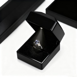 May include: A black ring box with an internal light showcasing a silver ring with a large blue gemstone and smaller side stones. The ring is set against a black velvet interior. Two other black boxes are visible.