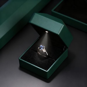 May include: A silver ring with a large blue gemstone and smaller white stones is displayed in an open, dark green ring box. The box has a built-in light shining on the ring, highlighting its details. The box is set against a dark background.